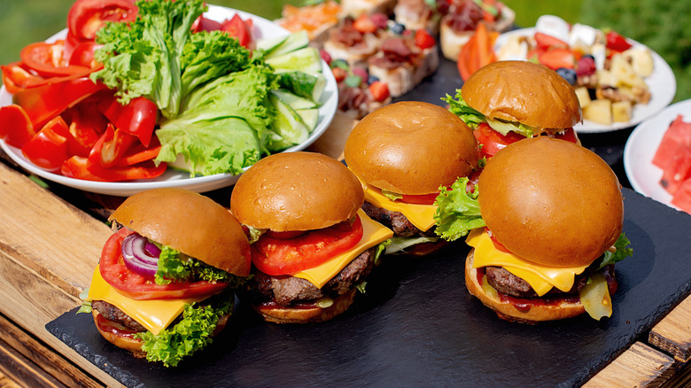 Cheeseburgers on an outdoor picnic table in front of a spread of food and a plate of peppers and lettuce