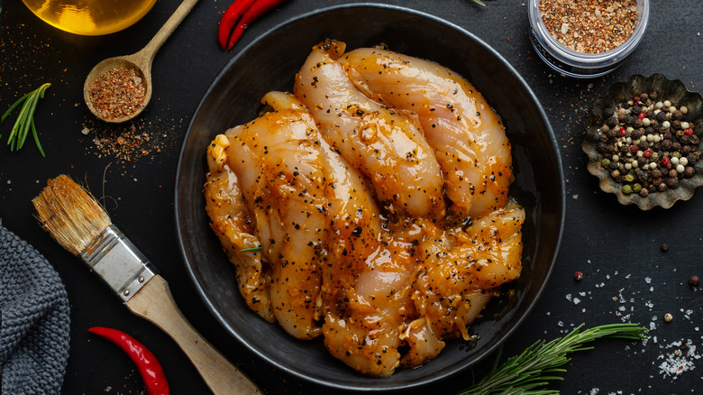 Raw marinated chicken breasts in bowl of marinade, surrounded by spices