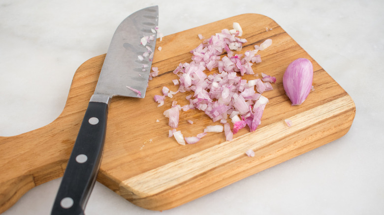 A minced shallot on a wooden cutting board with a knife and whole clove