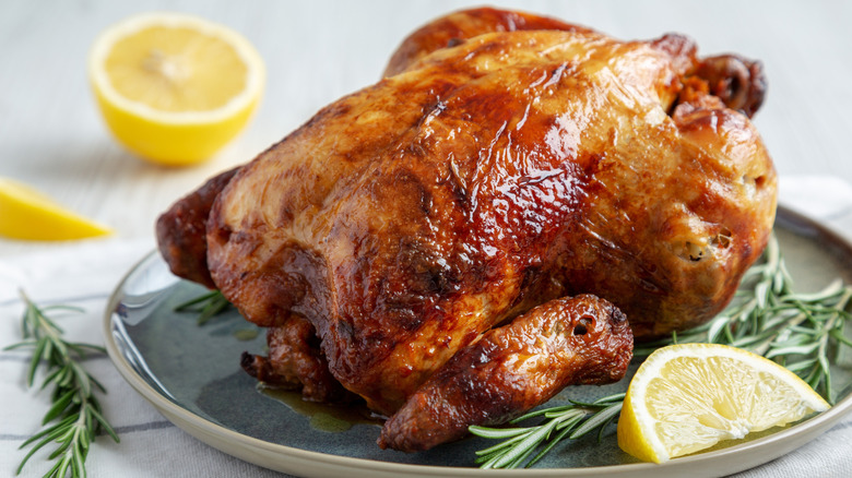 A whole, cooked rotisserie chicken on a plate with rosemary and a sliced lemon