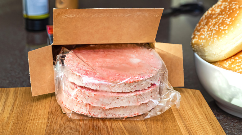 A box of frozen burger pattys on a wooden cutting board next ot a bowl of buns