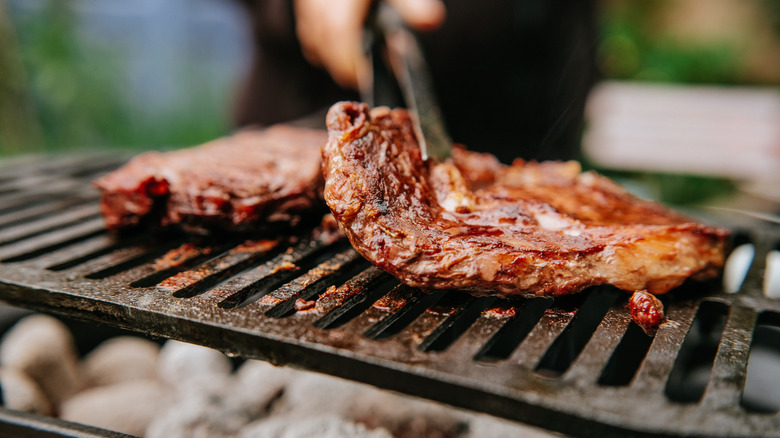 A person's hand grilling two steaks over charcoal