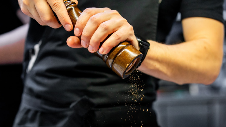 A person in a black apron is grinding pepper with a wooden grinder, capturing the dynamic motion of pepper flakes falling.