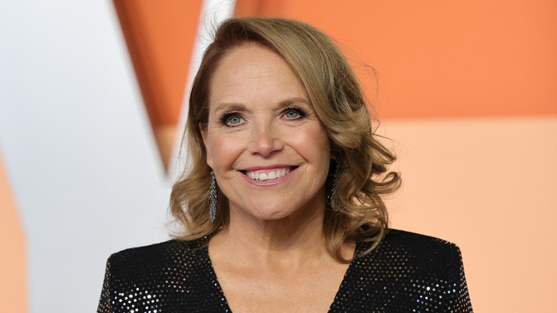 Katie Couric in a black dress in front of an orange background with white lettering