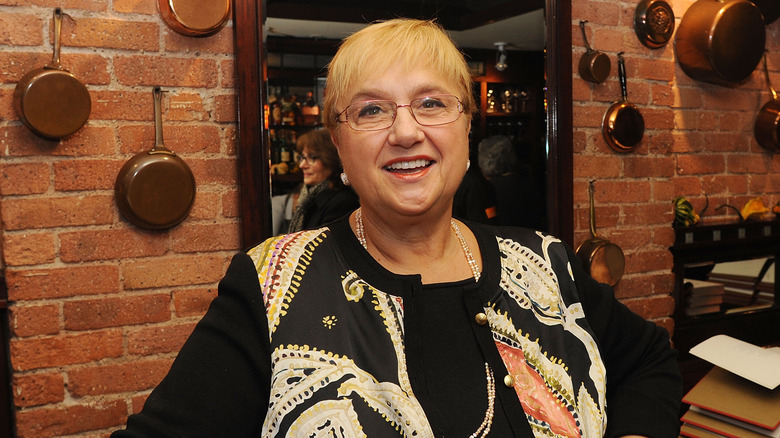 Lidia Bastianich wearing a black decorative shirt in front of a brick wall with copper pots and pans