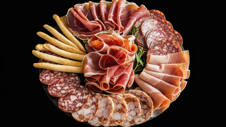 Deli meat platter with breadsticks on a black background.