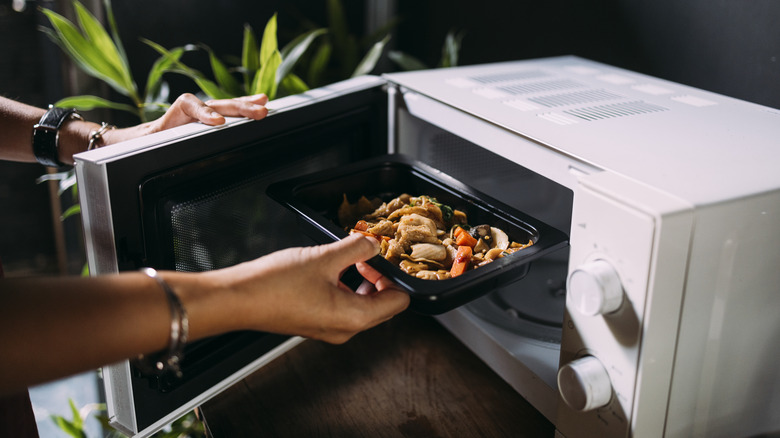 a person's hands putting a black takeout container with leftover food into a white microwave