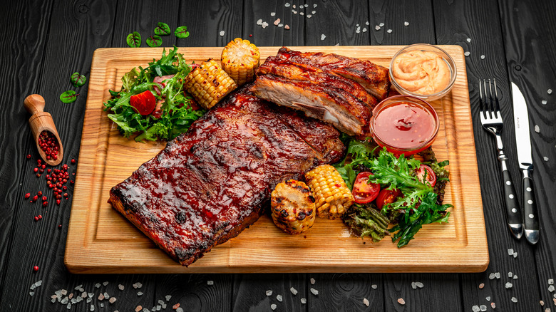 A rack of ribs on a wooden cutting board with grilled corn and vegetables with dishes of sauce