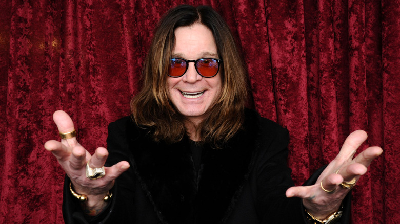 Ozzy Osbourne with his hands extended standing in front of a red velvet curtain