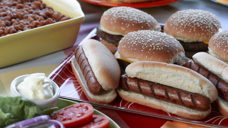 Hot dogs and burgers on buns with beans in the background and veggies on the side.