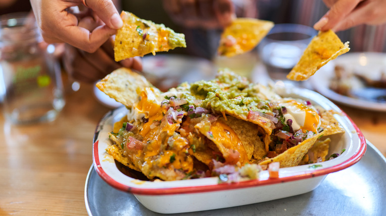 Hands grabbing from a dish of nachos on a wood table.