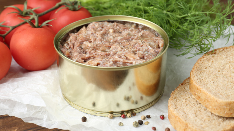 Canned meat on a wooden board with tomatoes on the vine, bread, peppercorns, and greens