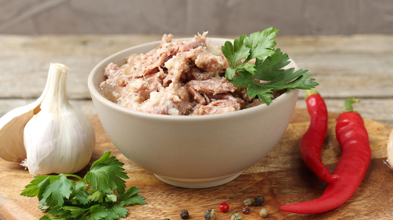 Canned meat in a bowl on a wooden cutting board with greens, garlic, peppers, and peppercorns