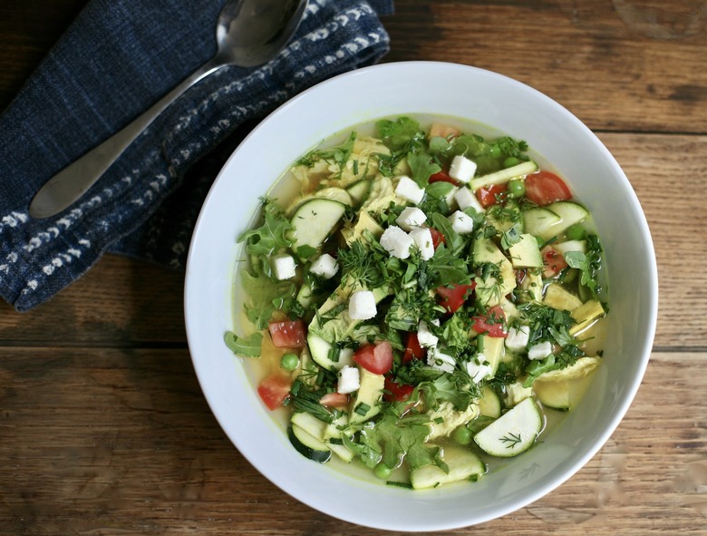 Spring chicken and greens soup recipe - photo of a white bowl on wood background; bowl is full of soup and greens, zucchini, tomato and topped with fresh herbs and fresh queso fresco