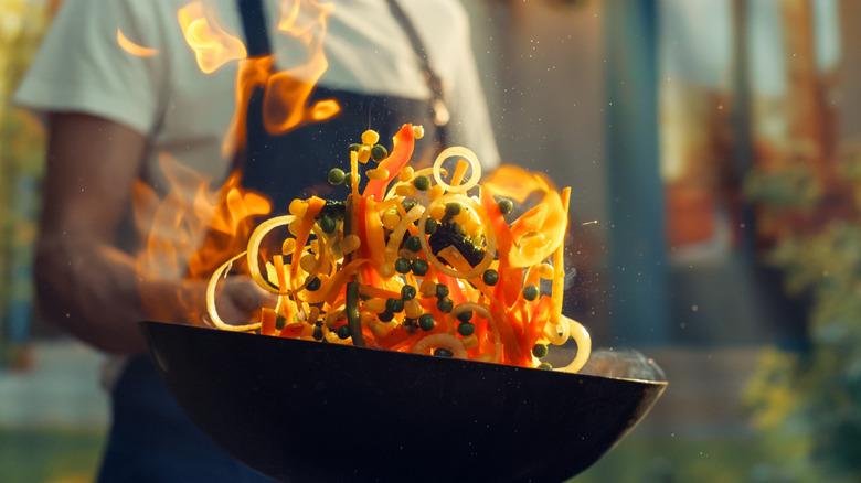 A chef holding a flaming wok tossing vegetables in a stir fry