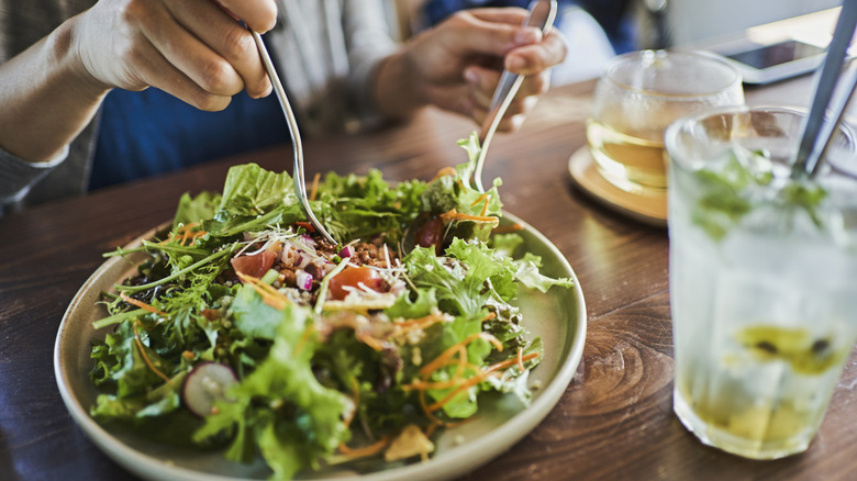 A person eating a salad with a cold drink in front of them on a wooden table