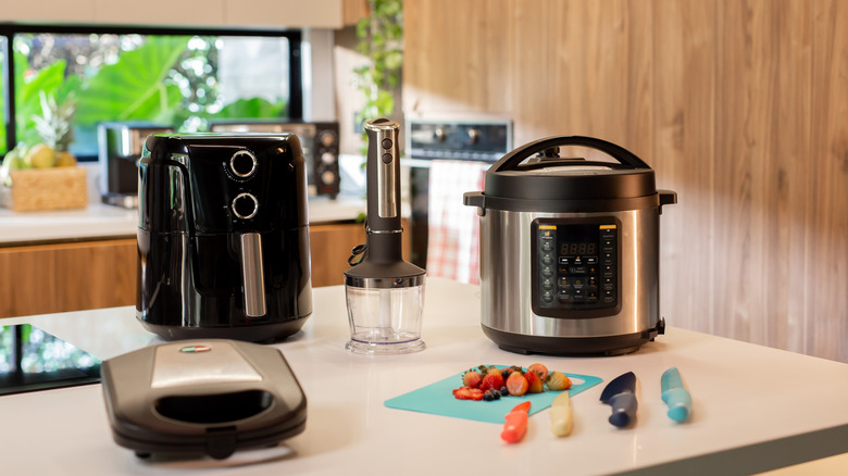 An air fryer, pressure cooker, blender and portable grill on a counter with some knives and sliced strawberries