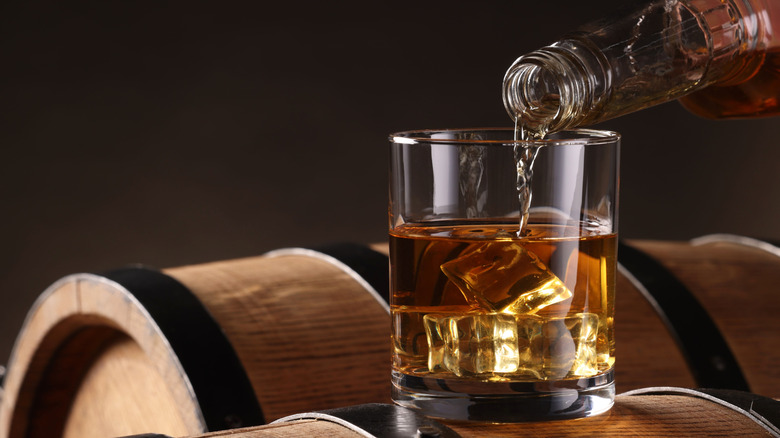 Glass of whisky being poured from a bottle on an aging barrel