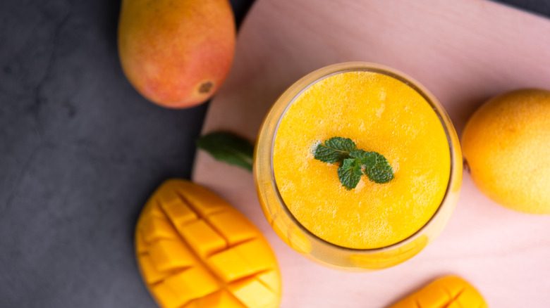 Top view of glass of mango juice garnished with mint leaves, surrounded by mangoes