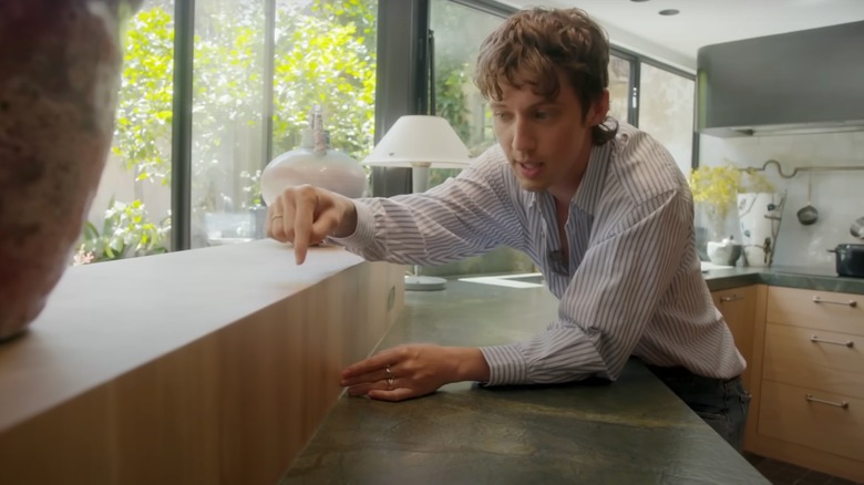Troye Sivan points to his wooden counter, while standing in his kitchen