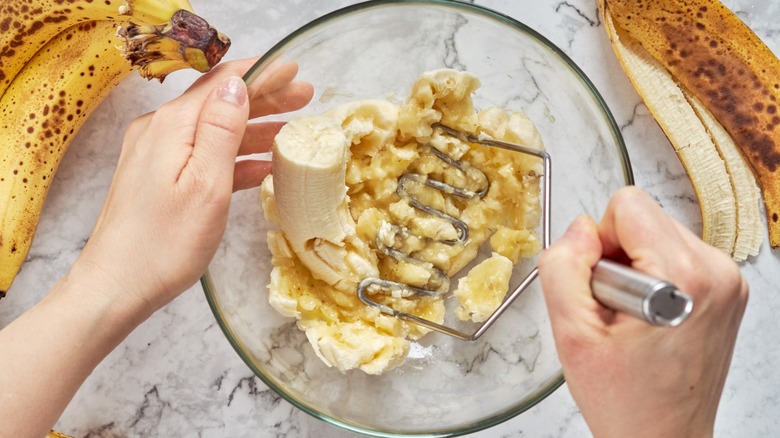 Person using a potato masher to mash bananas in a glass mixing bowl with bananas and peels on the counter
