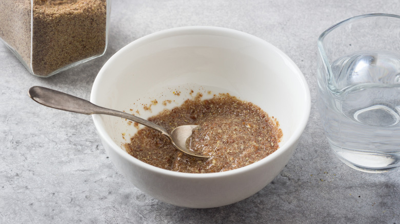 White bowl of soaked flaxseed with a spoon and water and ground flaxseed on a counter