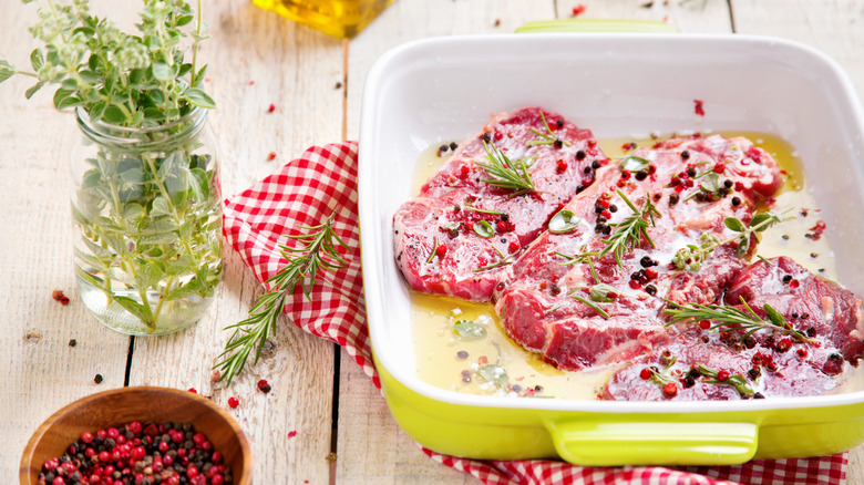 steaks marinating in a dish surrounded by herbs and a bottle of olive oil