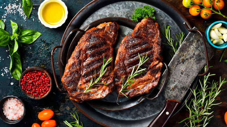 Two grilled steaks in a cast iron pan surrounded by various herbs and aromatics