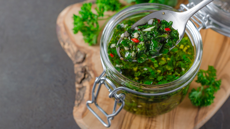 Spooning out some homemade chimichurri sauce from a jar on a wood board with herbs