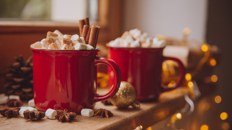 Two red mugs of hot chocolate with marshmallows and cinnamon sticks