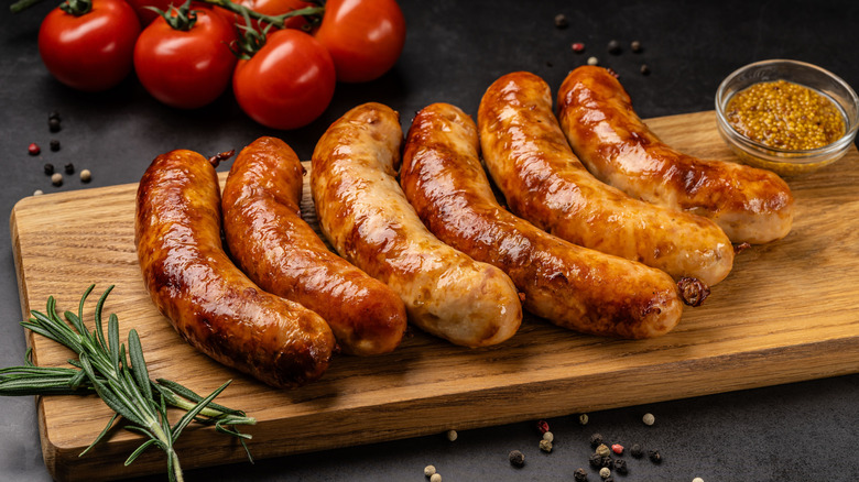 Six plump, browned sausages on a cutting board with mustard seed, rosemary, and tomatoes