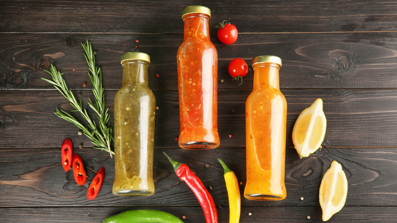 Three jars of hot sauce laying on a dark wood surface with lemon wedges, cherry tomatoes, chilies, herbs, and peppercorns