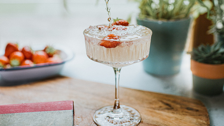 Clear beverage in a tall-stemmed glass with strawberry garnish on a wooden table with plants and strawberries in the background..