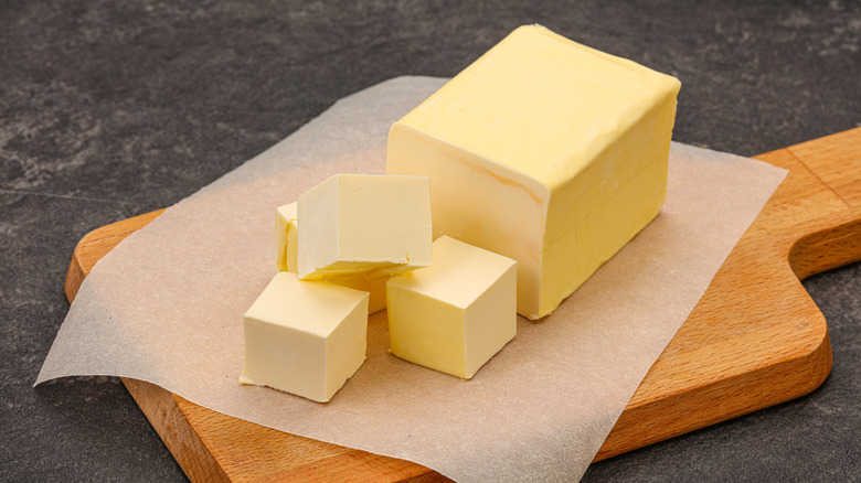 A stick of butter partially broken into cubes, resting on parchment paper on a brown wooden cutting board