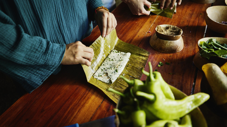 Person making a tamale in a corn husk on a wood table with peppers in the forefront.