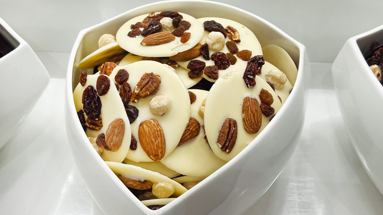 A shield-shaped white bowl filled with white chocolate discs, topped with almonds, raisins, and dried cranberries