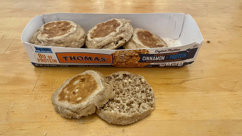 A container of Thomas' cinnamon protein English muffins with one cut in half in front of the box