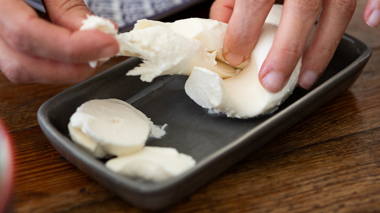 Close up of hands tearing Bufala mozzarella cheese in a small tray on a wood table.