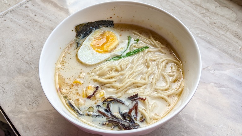 A bowl of creamy ramen with noodles with egg, corn, and seaweed on a table