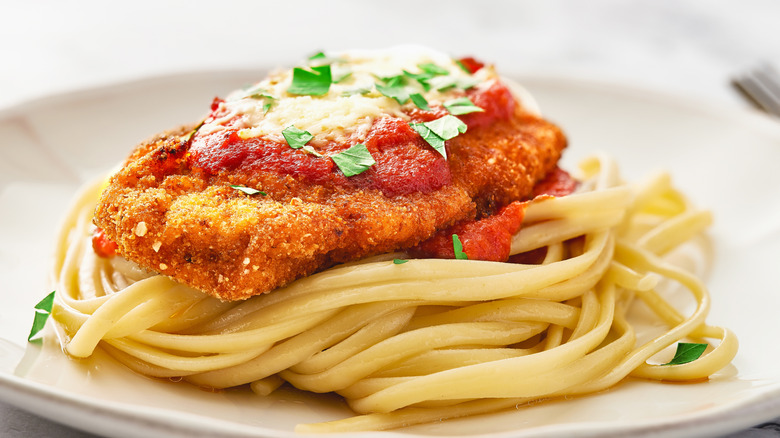 A fried chicken cutlet coated in breadcrumbs, topped with red sauce, white cheese, and green parsley, on a bed of fettuccine