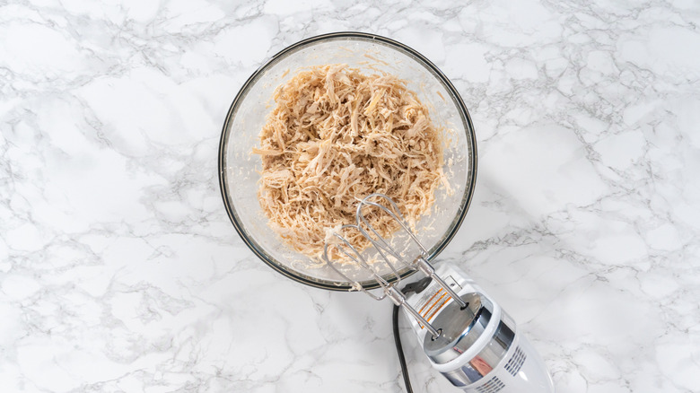 A bowl of shredded chicken with a hand mixer on a marble table