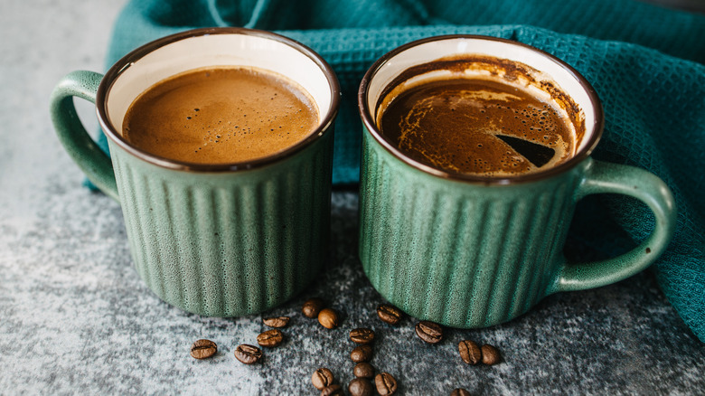 Two cups of coffee in green mugs with coffee beans in front and green towel in back