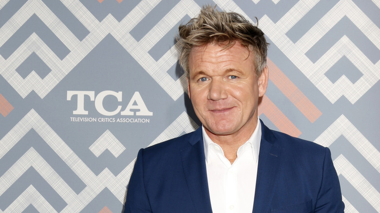 Gordon Ramsey in a navy blue suit and white suit smiling to camera.