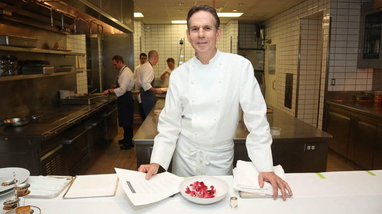 Thomas Keller in chef's whites standing in a kitchen smiling at the camera.