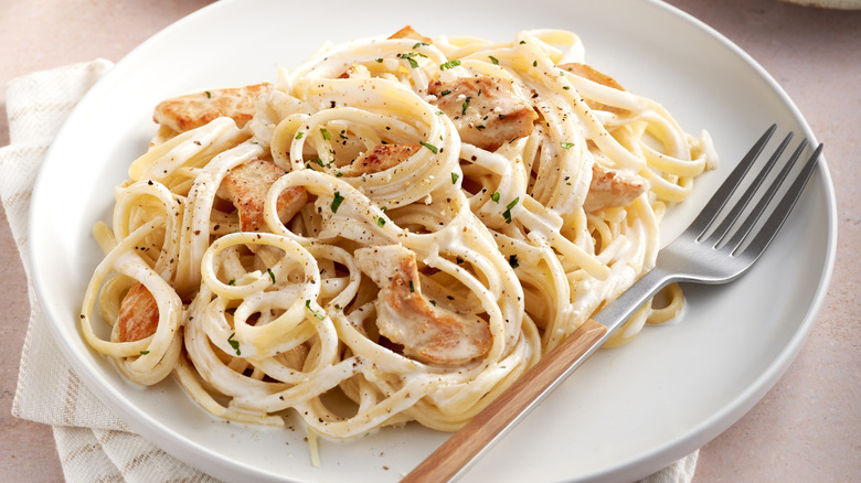 A white plate with chicken Alfredo pasta with seasonings and a metal fork on top of a cloth and table