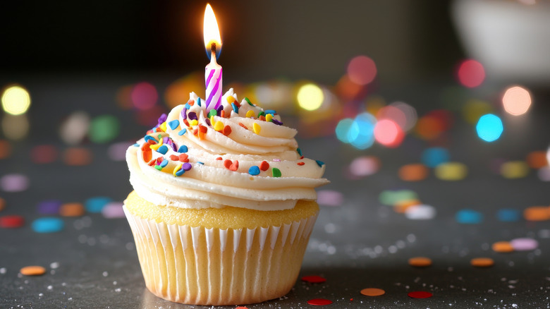 Vanilla cupcake with frosting, sprinkles, and a lit candle on top, against a blurred festive background