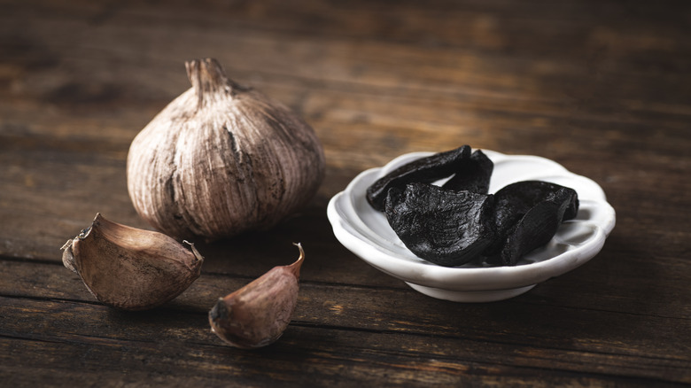 An unpeeled bulb of black garlic, with unpeeled loose cloves, and a bowl of peeled black cloves