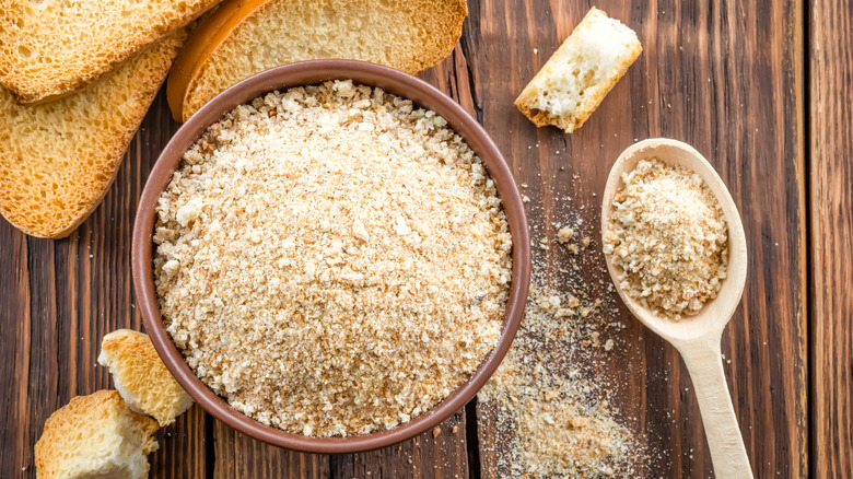 Wooden table with bread and a bowl of breadcrumbs with a large spoon of breadcrumbs.