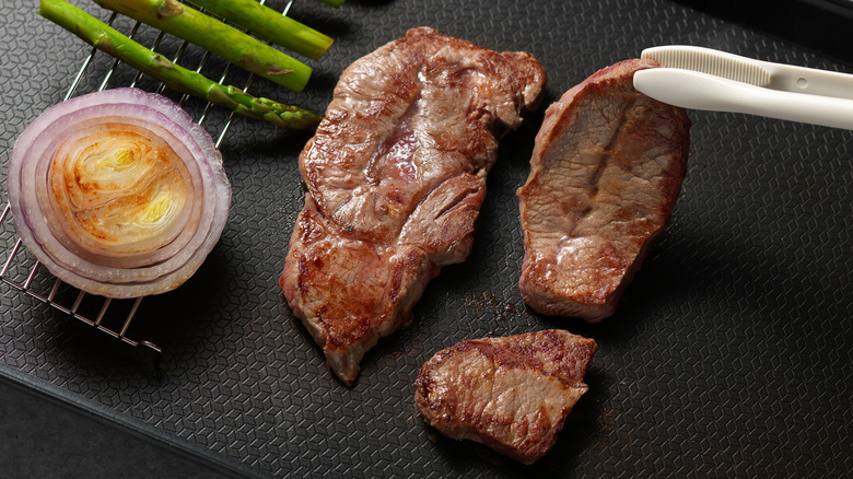 Tongs picking up seared Kobe beef next to grilled onion and asparagus