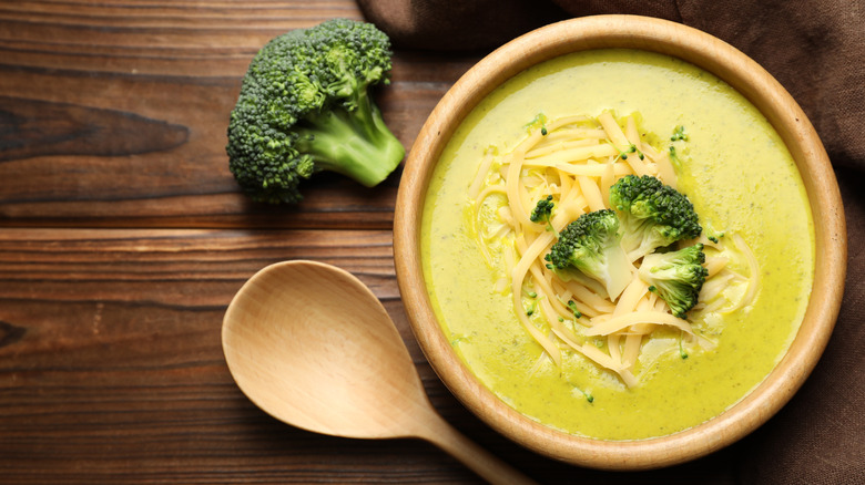 ﻿﻿A wooden bowl of broccoli cheese soup topped with broccoli cuts and shredded cheese